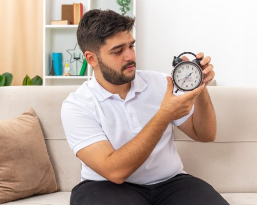 young-man-casual-clothes-holding-alarm-clock-looking-it-with-serious-face-sitting-couch-light-living-room_141793-98977